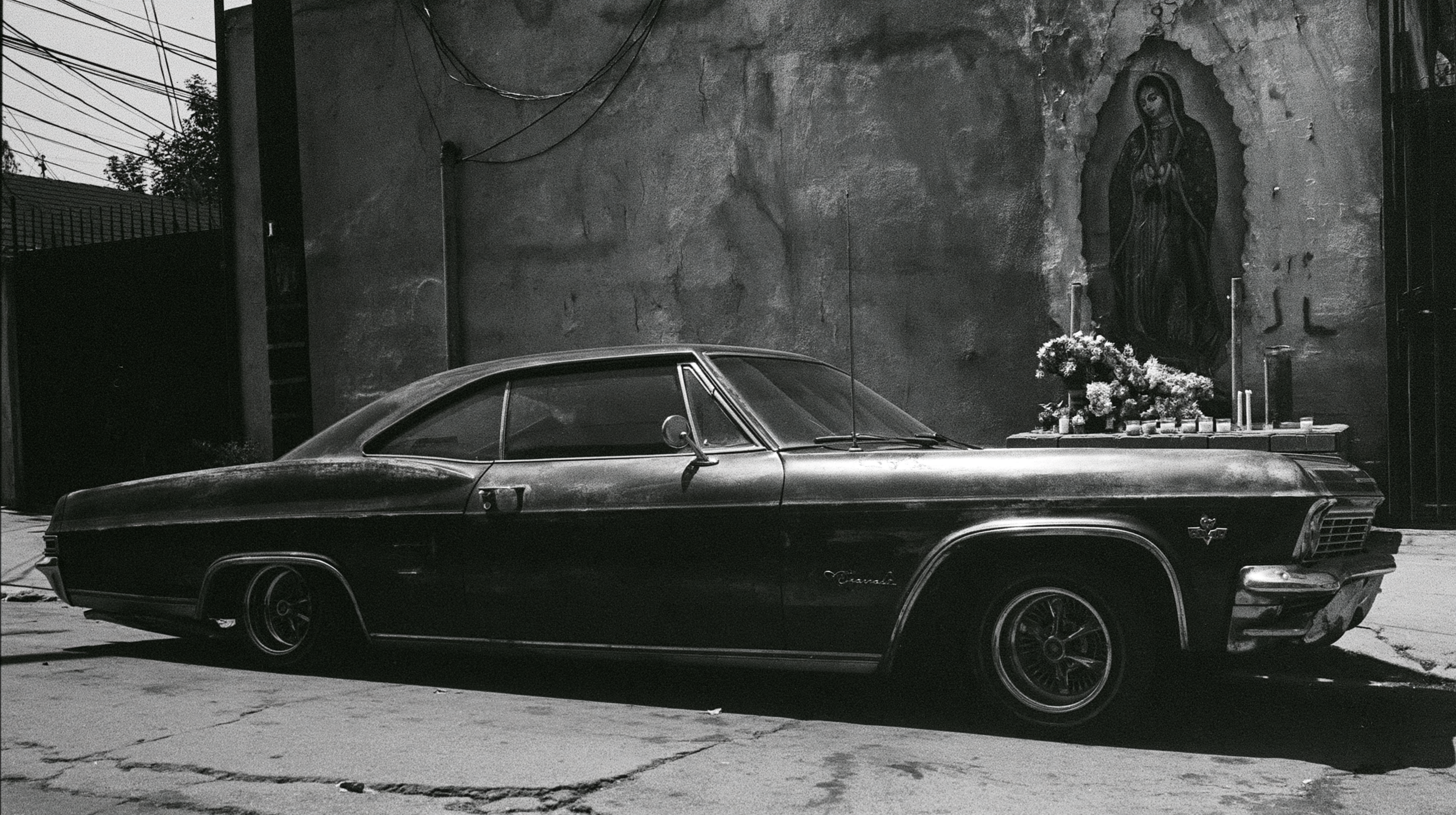 A 1960s Chevrolet Impala lowrider parked on a sun-drenched East Los Angeles street with a weathered Virgin of Guadalupe shrine visible on the stucco wall behind it, captured in black and white 35mm documentary photography style.