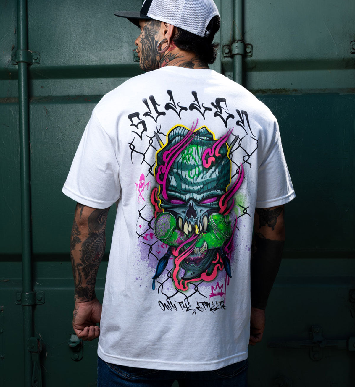 Person wearing a white t-shirt with a colorful graphic design on the back, standing against a green wall.
