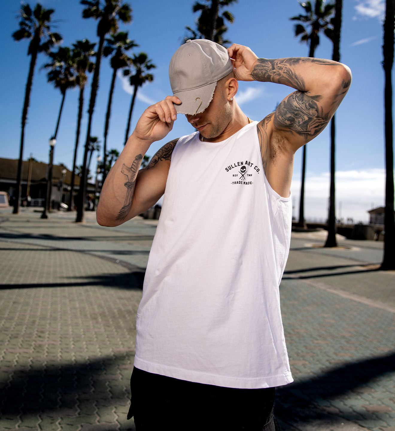 Man wearing a white tank top with a logo, standing on a sidewalk with palm trees in the background.
