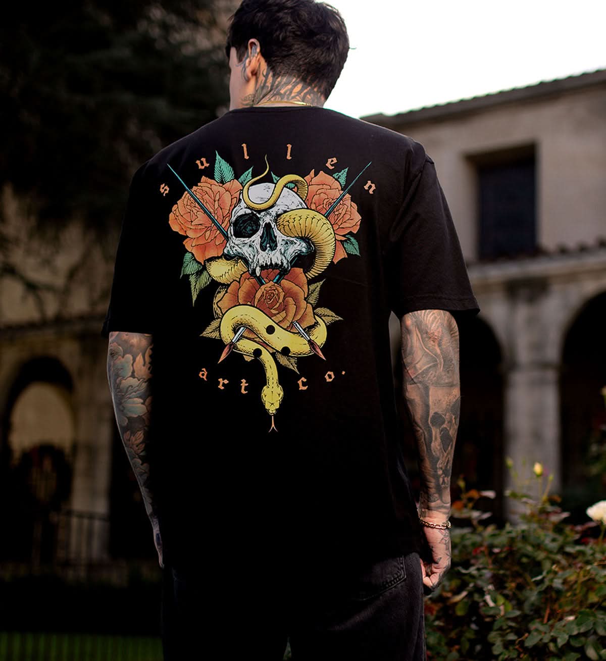 Person wearing a black t-shirt with a colorful graphic design featuring a skull, roses, and a snake, with 'Sullen Art Co.' branding, outdoors.