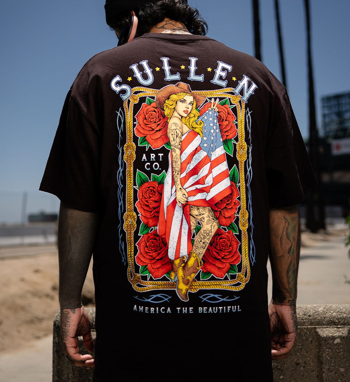 Person wearing a black t-shirt with a colorful graphic design featuring an American flag and roses, branded 'Sullen'.