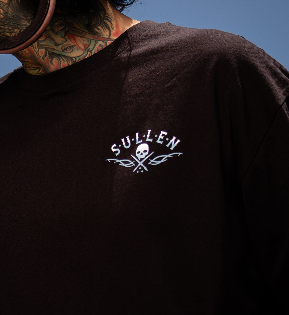 Black t-shirt with 'Sullen' logo worn by a person with tattoos against a blue sky.