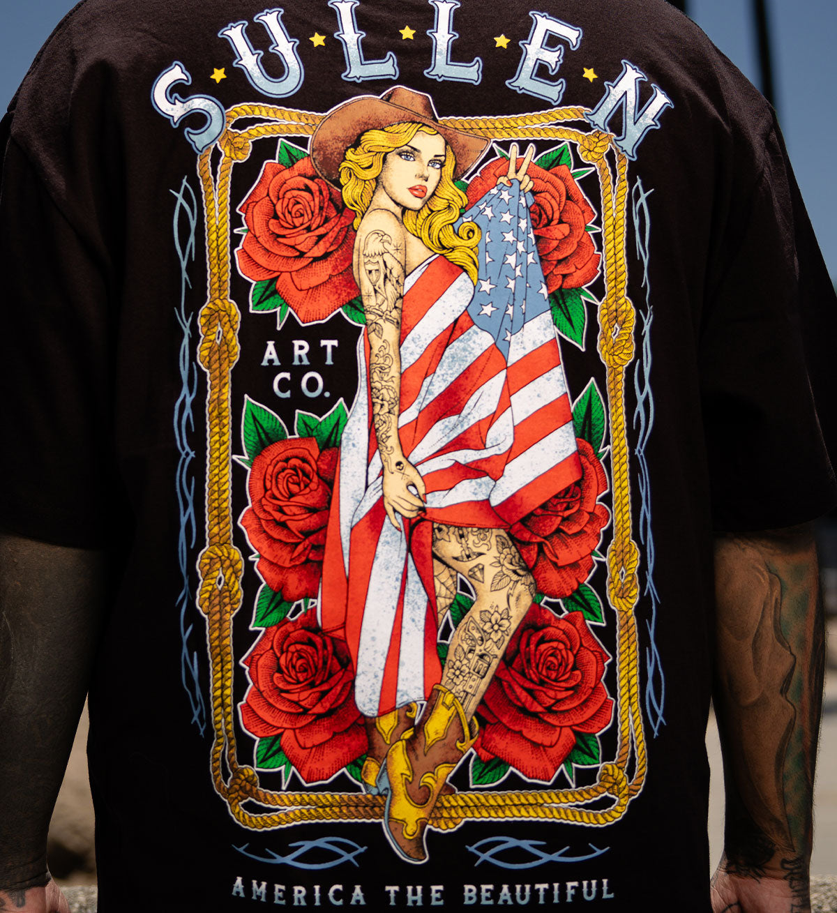 Black t-shirt with a colorful graphic of a woman holding an American flag surrounded by roses, branded 'Sullen Art Co'.