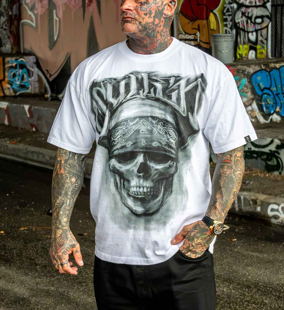 A tattooed man wearing a white graphic t-shirt with a large black and gray skull design on the front, standing in front of a graffiti-covered wall.