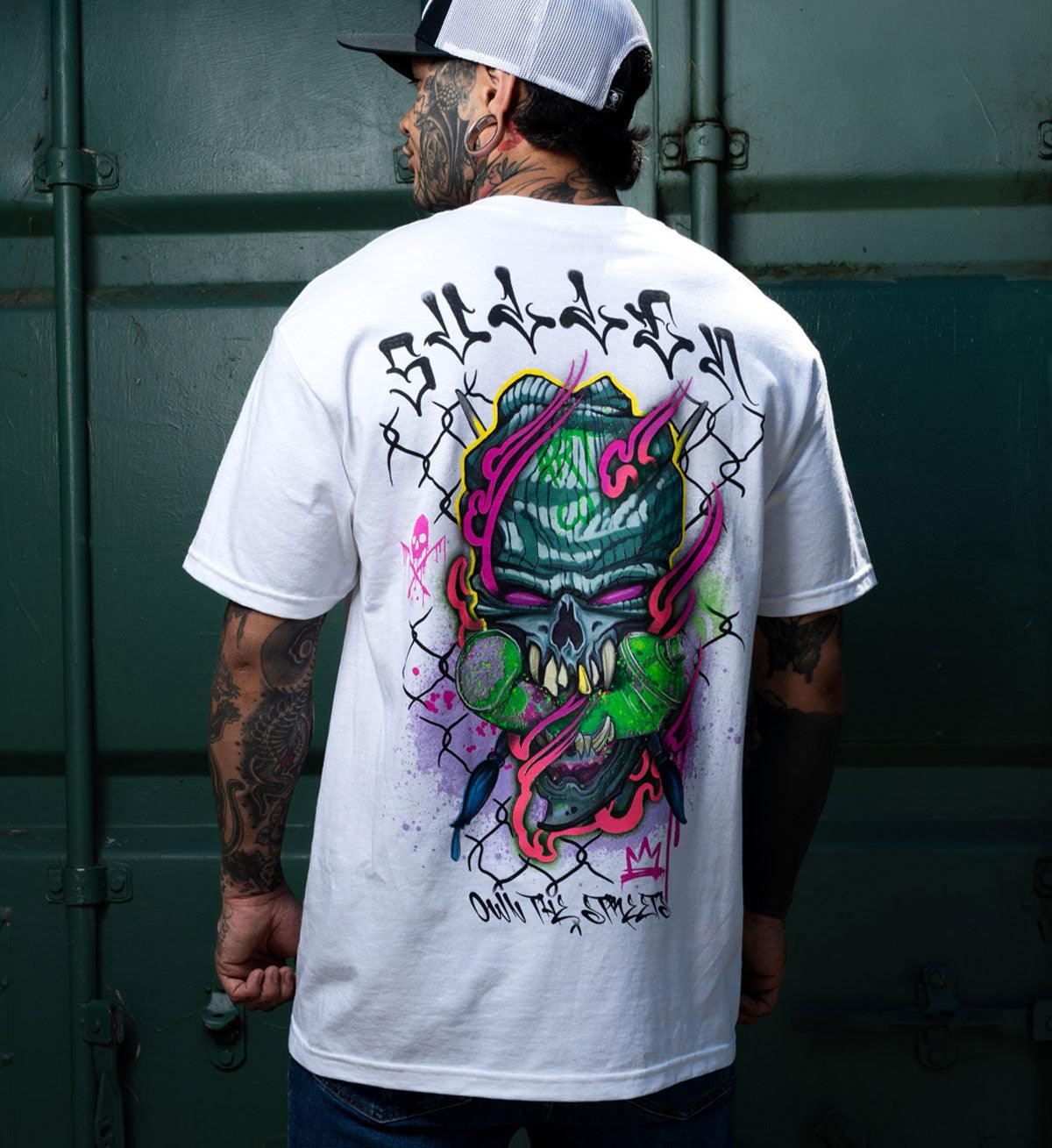 Person wearing a white t-shirt with a colorful graphic design on the back, standing against a green wall.