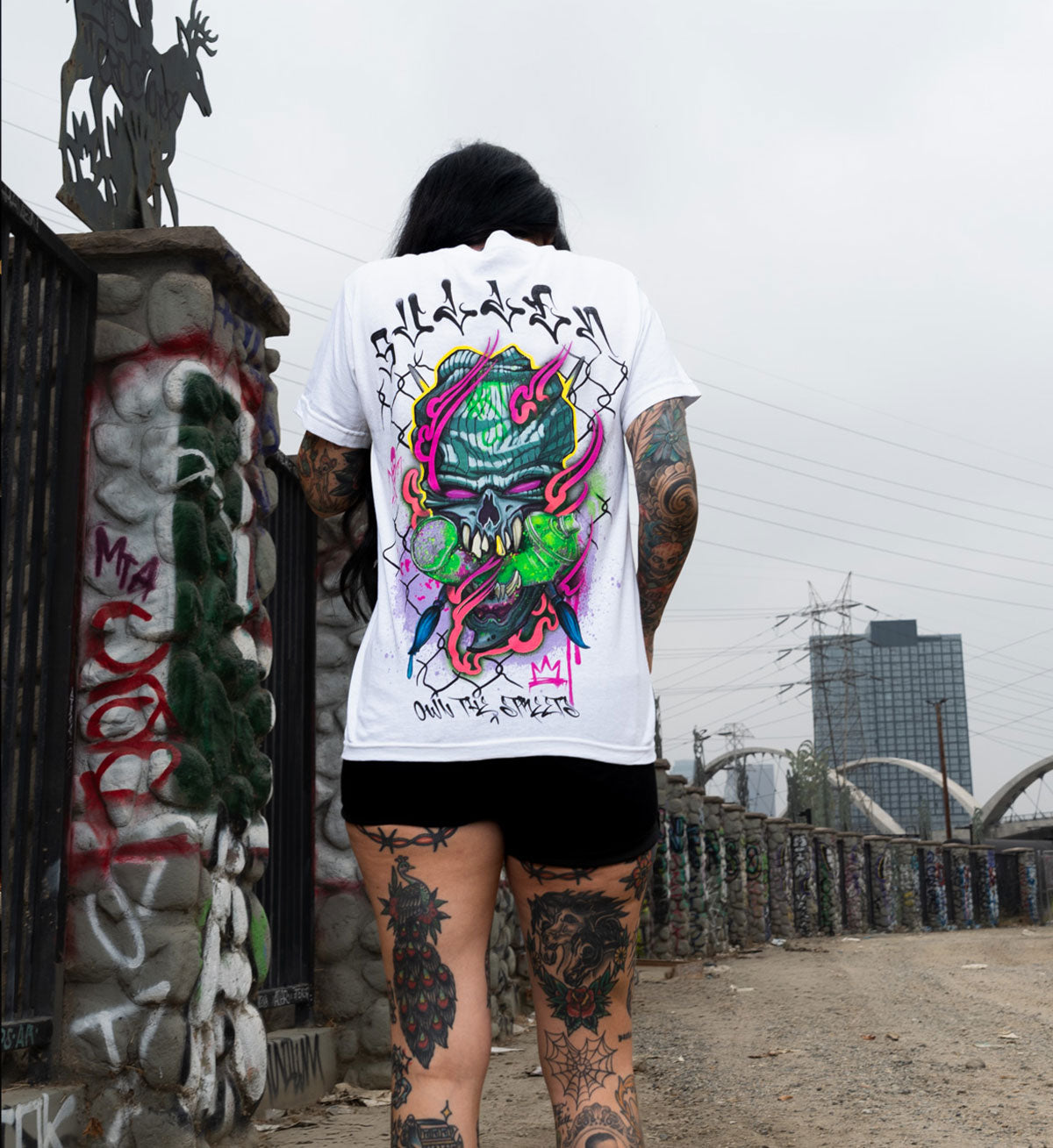 Person wearing a white t-shirt with colorful graphic design, standing in an urban setting with graffiti and a bridge.