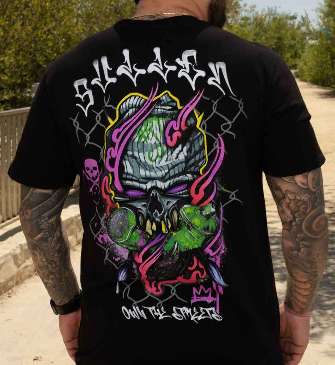 A man viewed from behind wearing a black t-shirt with a large colorful graphic on the back. The graphic features a skull, barbed wire, and the text 'Sudden' and 'own th streets' in white and neon colors.