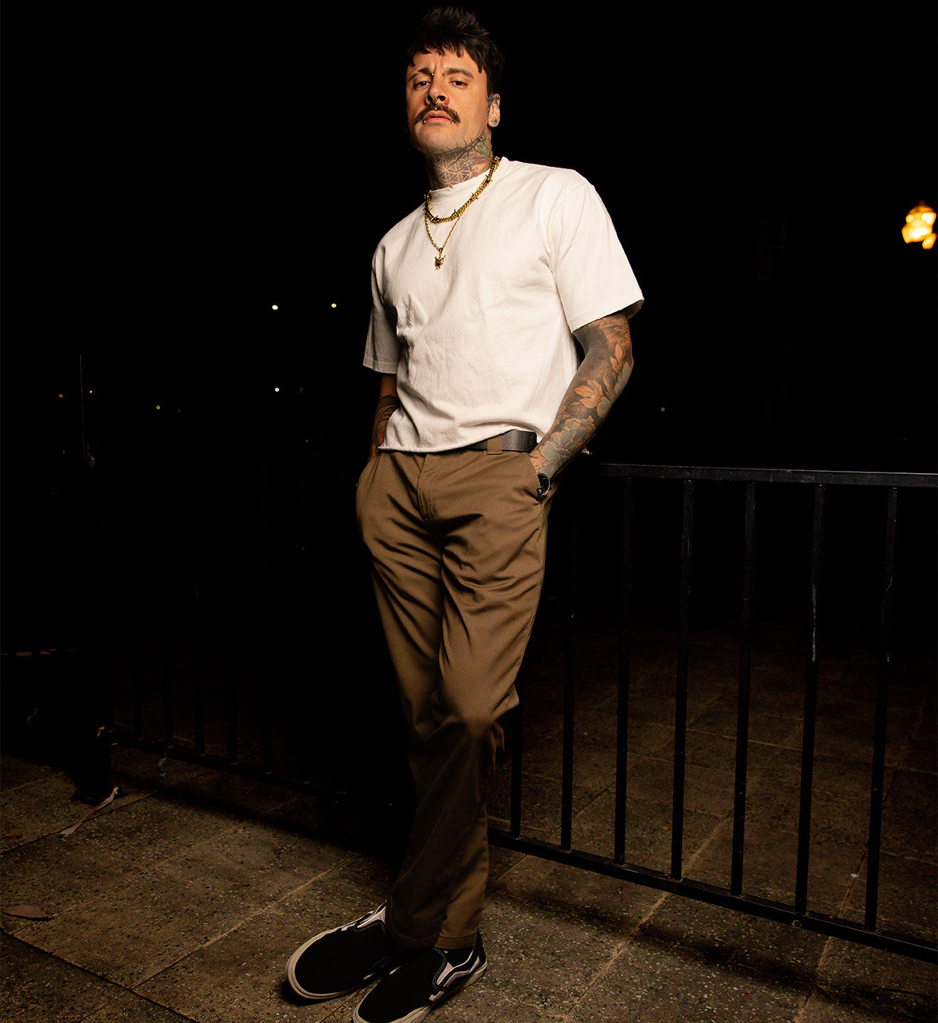 Man wearing a white t-shirt and brown pants standing against a dark background