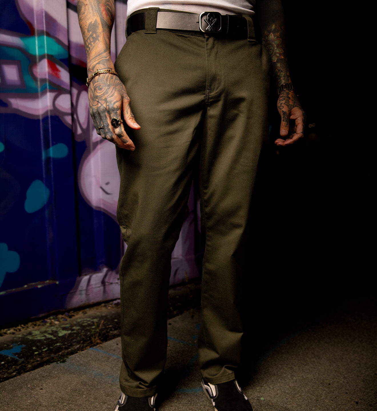 Person wearing olive green pants with tattoos on arms and hands, standing against a dark background.
