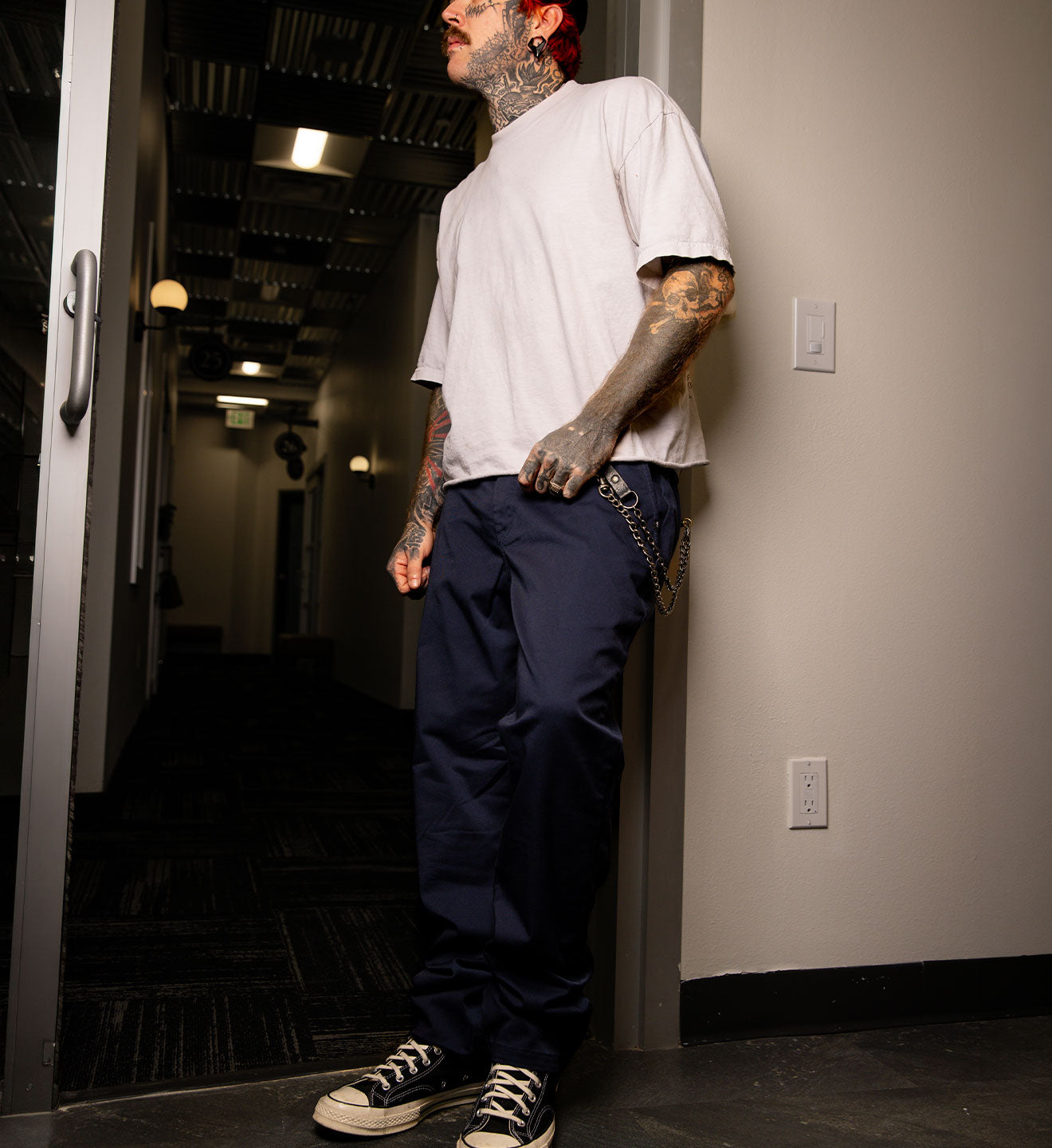 Person wearing a white t-shirt and blue pants leaning against a wall in a dimly lit hallway.