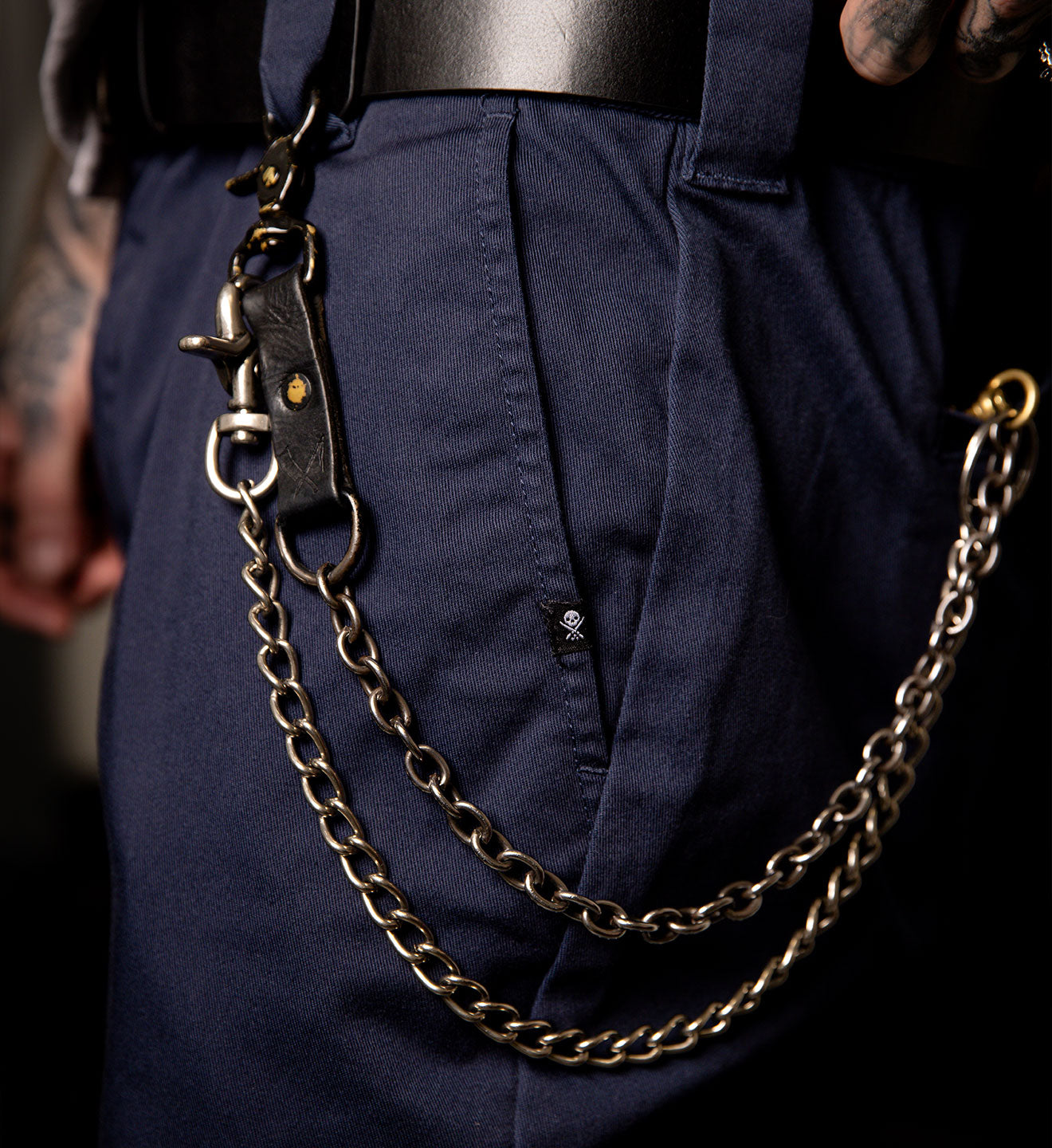 Gold chain hanging from a belt on navy blue pants with tattoos visible.