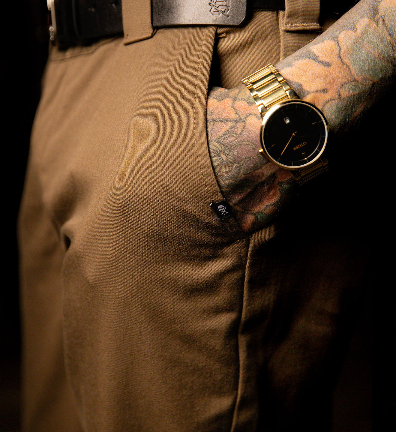 Gold watch on a tattooed wrist with brown pants and belt.
