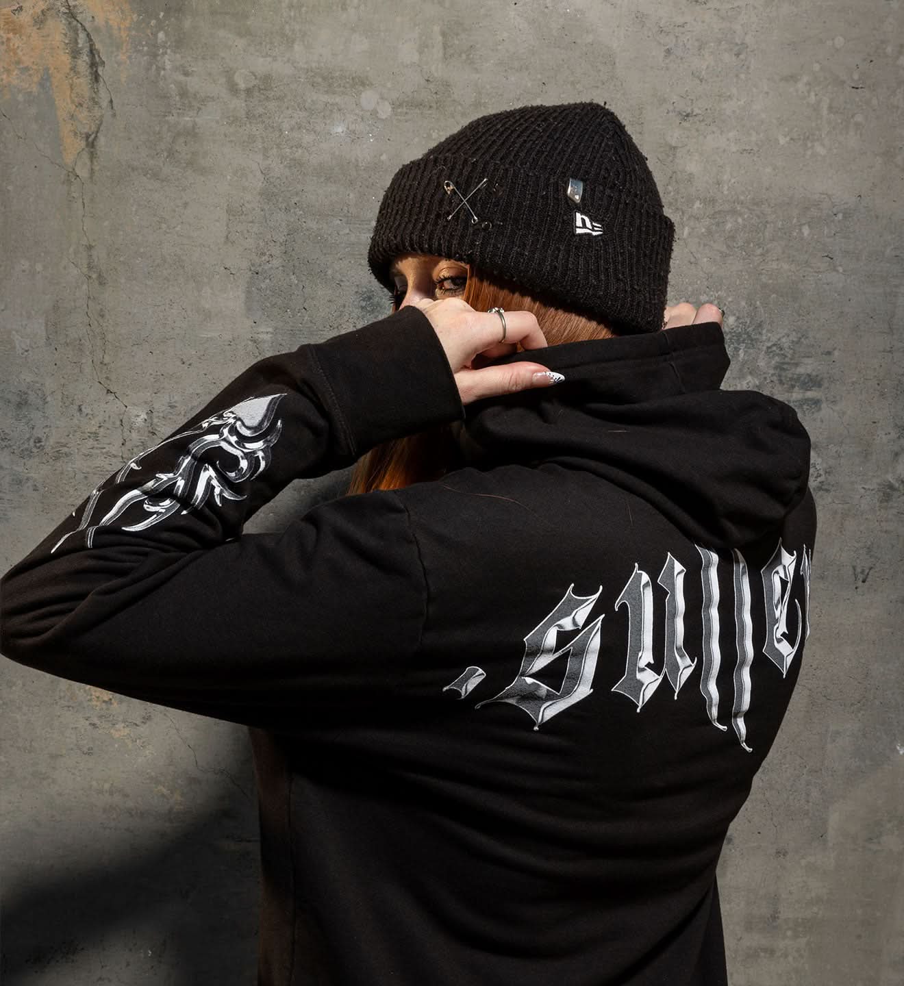Person wearing a black hoodie with 'Sullen' branding, standing against a textured gray wall.