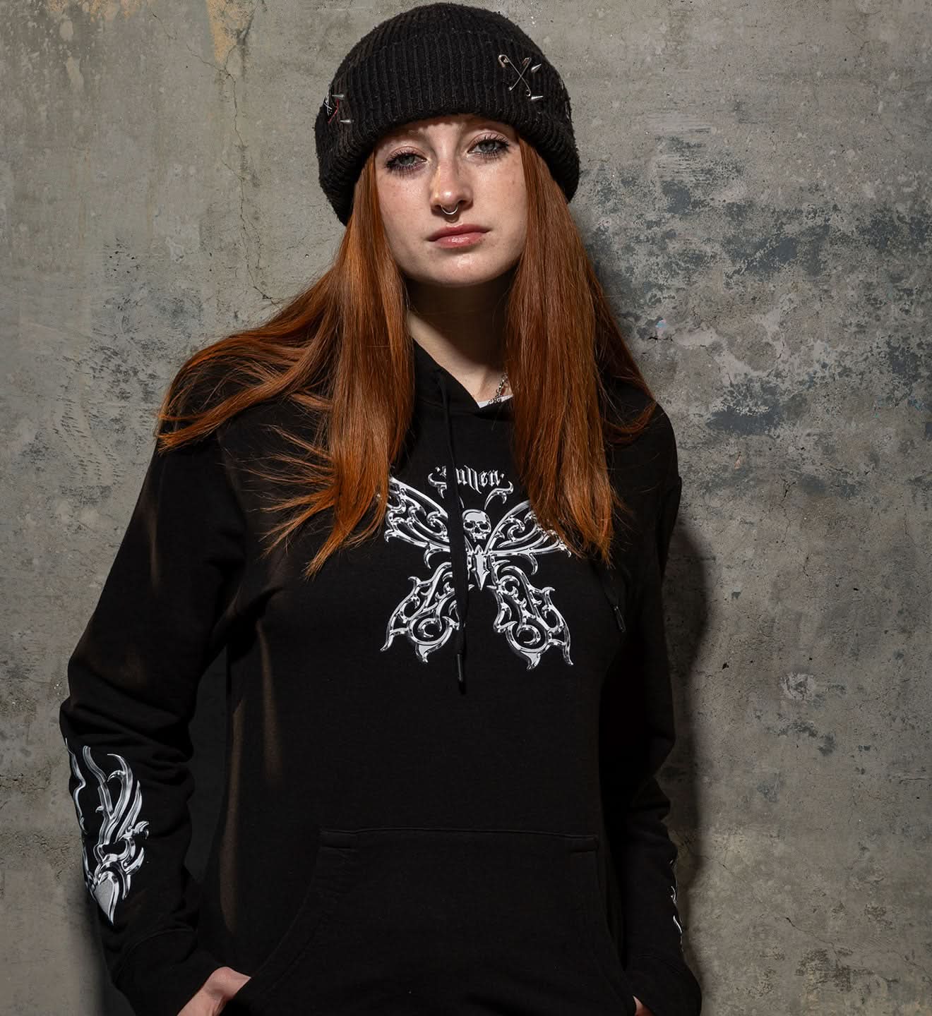 Person wearing a black hoodie with white designs and a beanie against a textured gray wall.
