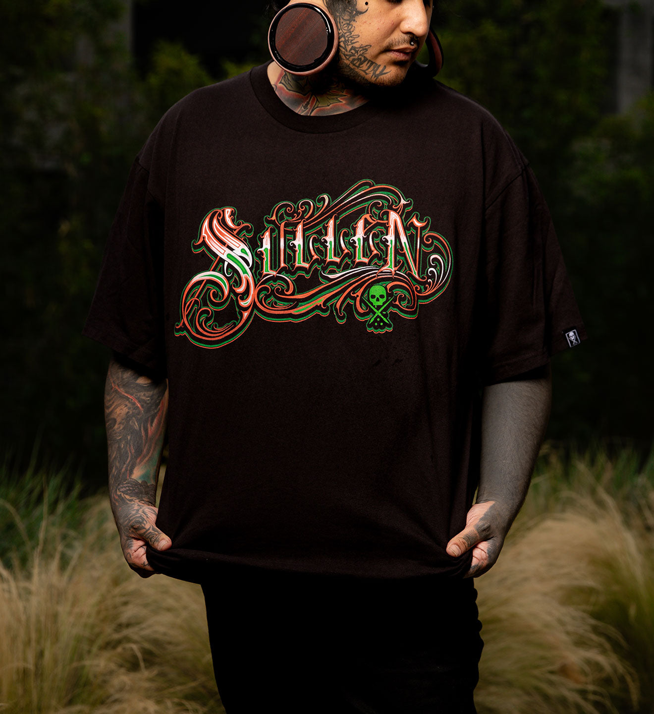 Person wearing a black t-shirt with colorful 'Sullen' logo in a natural setting