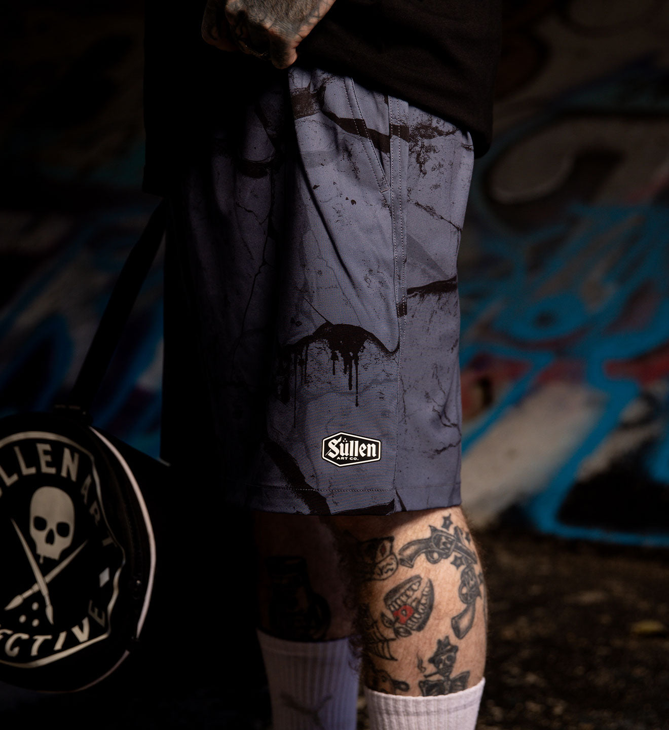 Person wearing a gray camouflage jacket with visible tattoos, holding a product with 'Sullen' branding.
