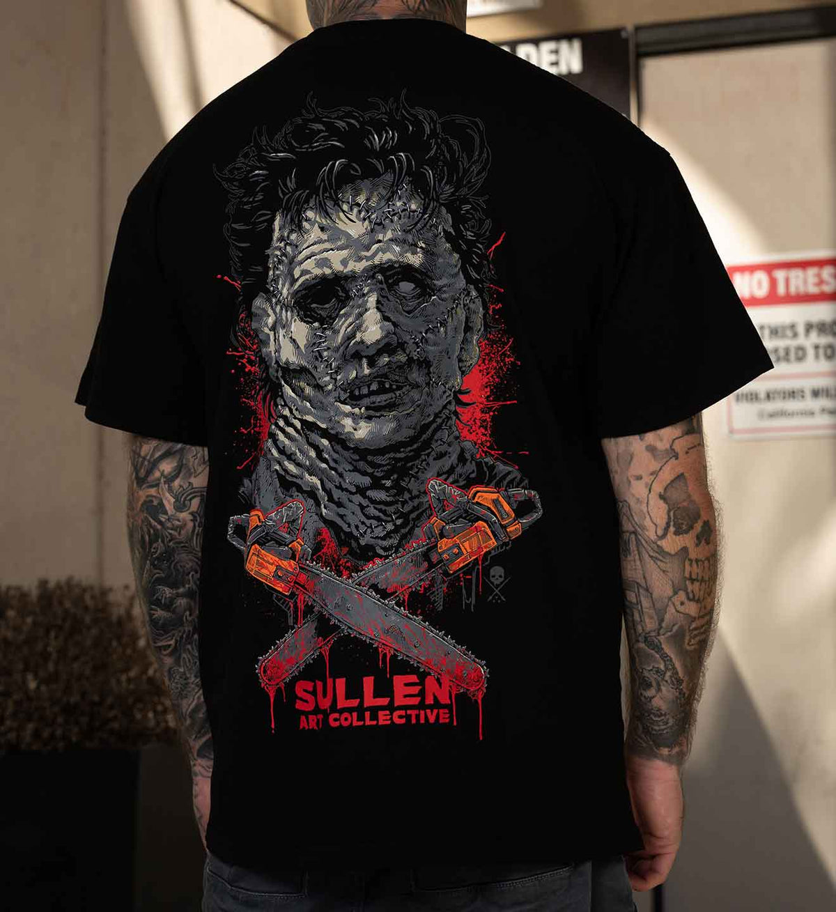 A man viewed from behind wearing a black graphic t-shirt featuring a horror character design with chainsaws and the text 'SULLEN ART COLLECTIVE'.