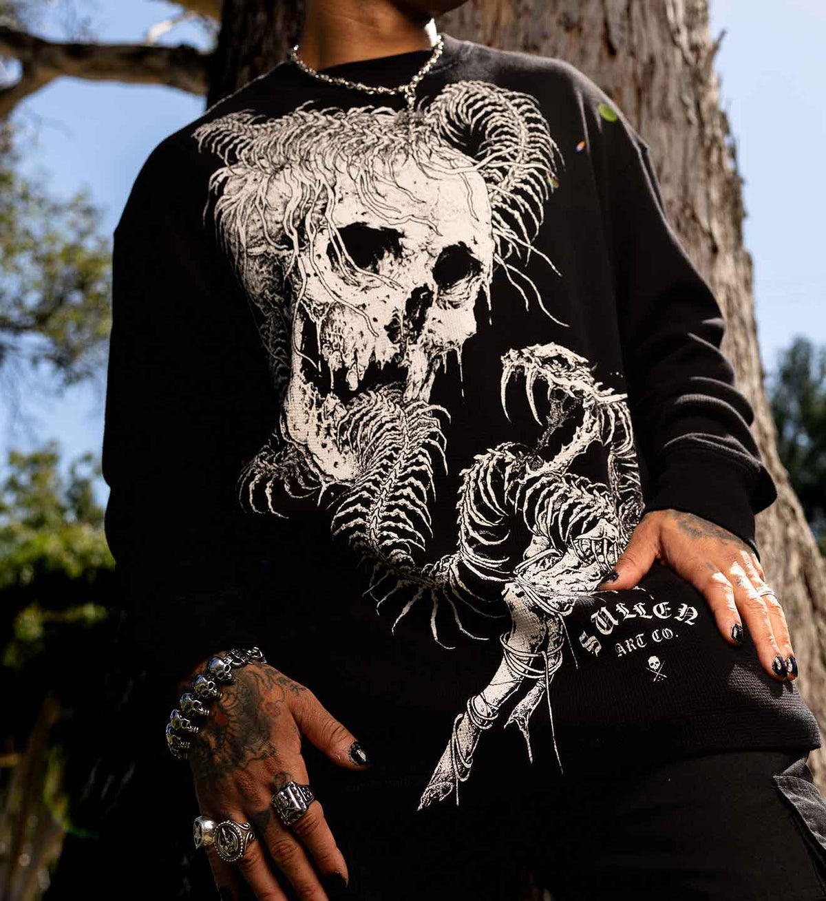 A person outdoors wearing a black crewneck sweatshirt with a large white skeletal skull and snake graphic, paired with a black beanie featuring white gothic text.