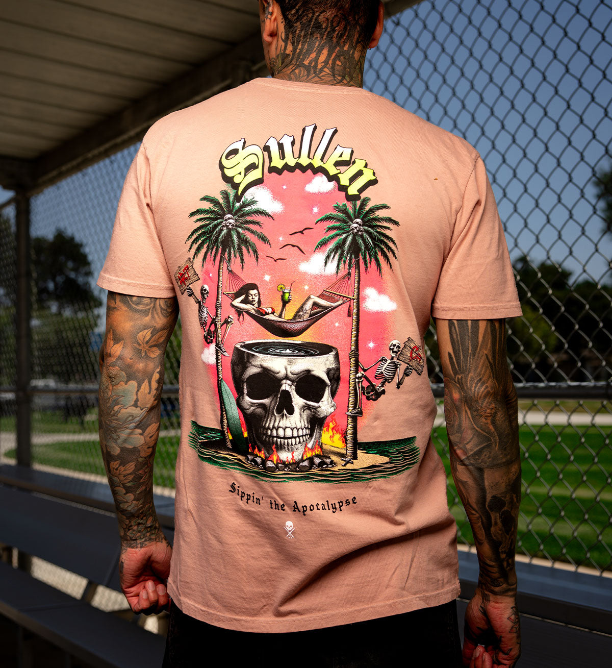 Person wearing a pink t-shirt with a graphic design featuring a skull, palm trees, and text.
