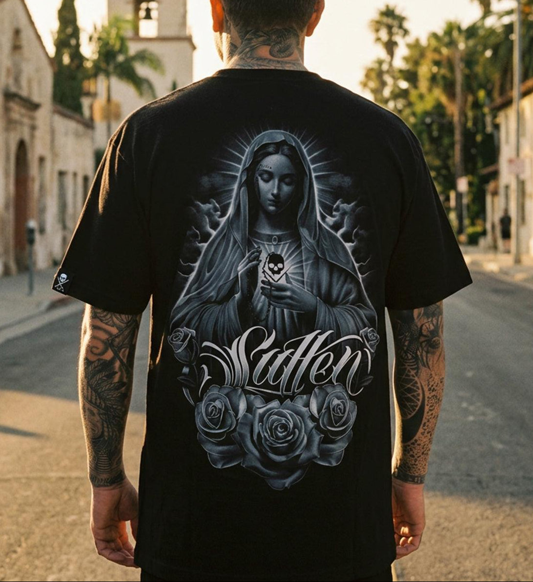 A man viewed from behind wearing a black t-shirt with a large graphic of the Virgin Mary and the word 'Suffer' on the back, standing on a sunlit street with buildings and palm trees in the background.