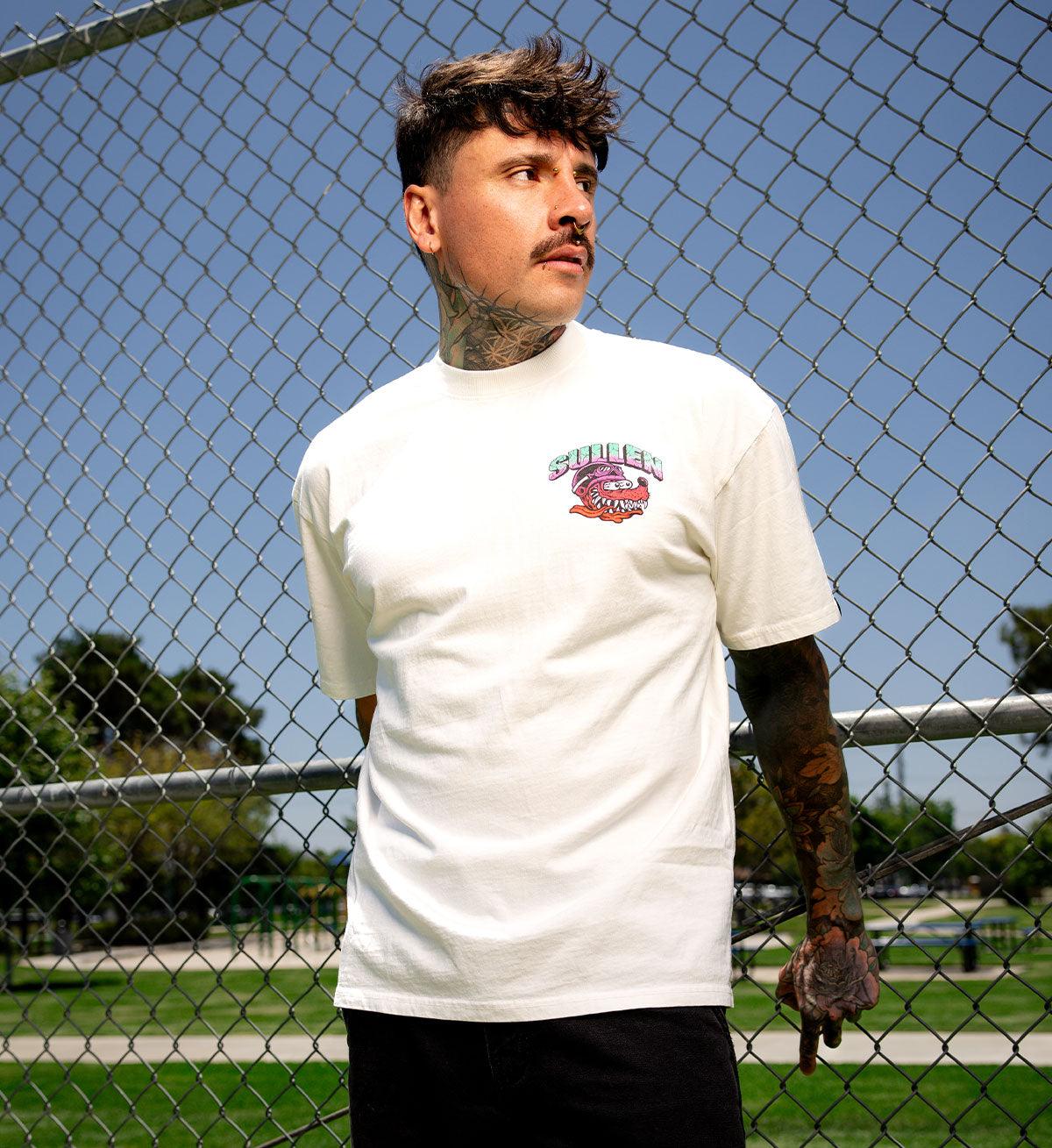 Man wearing a white t-shirt with a logo in front of a chain-link fence.