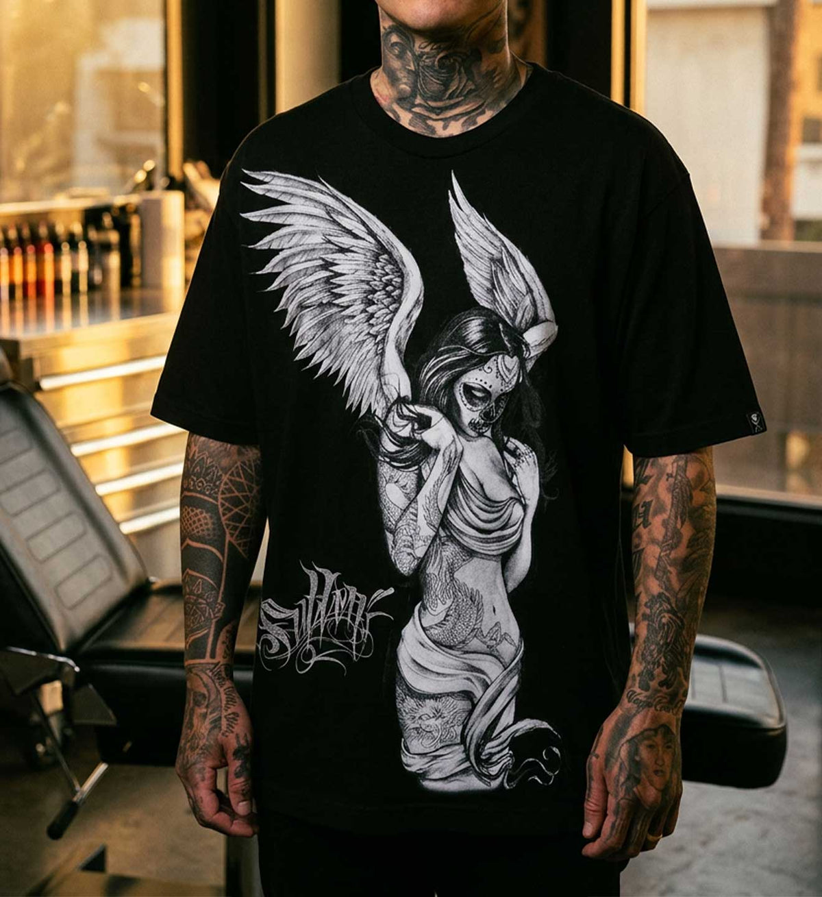 A tattooed man wearing a black graphic t-shirt with a detailed black and white illustration of an fallen angel and the word 'SULLEN' on the front, set against a blurred tattoo studio background.