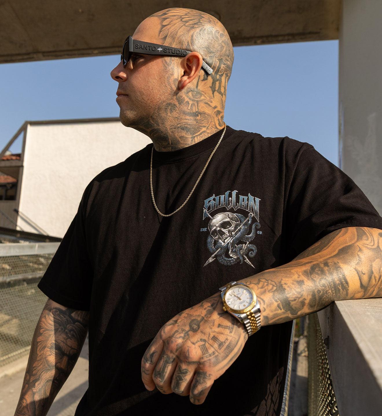 Man with tattoos wearing a black t-shirt with a graphic design, gold watch, and sunglasses on a rooftop.