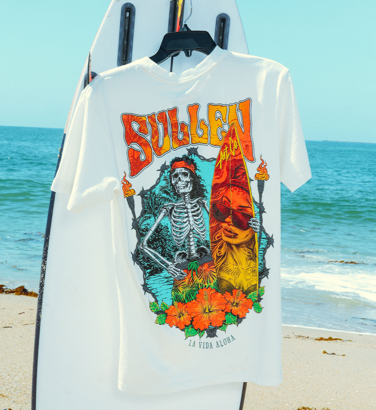 White t-shirt with colorful graphic design featuring a skeleton and flowers, displayed on a beach.