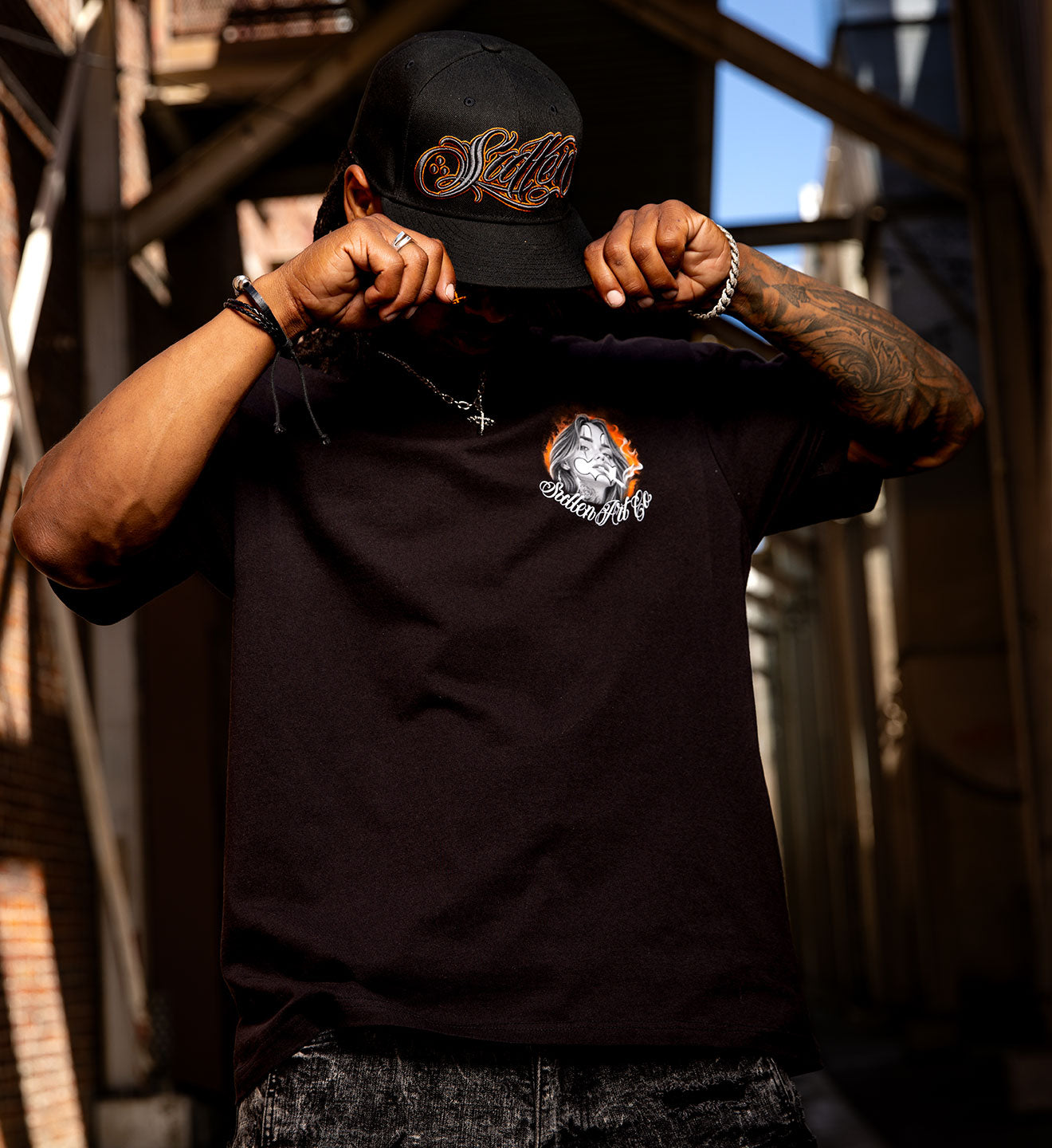 Person wearing a black t-shirt and cap with logos, standing in an industrial setting.