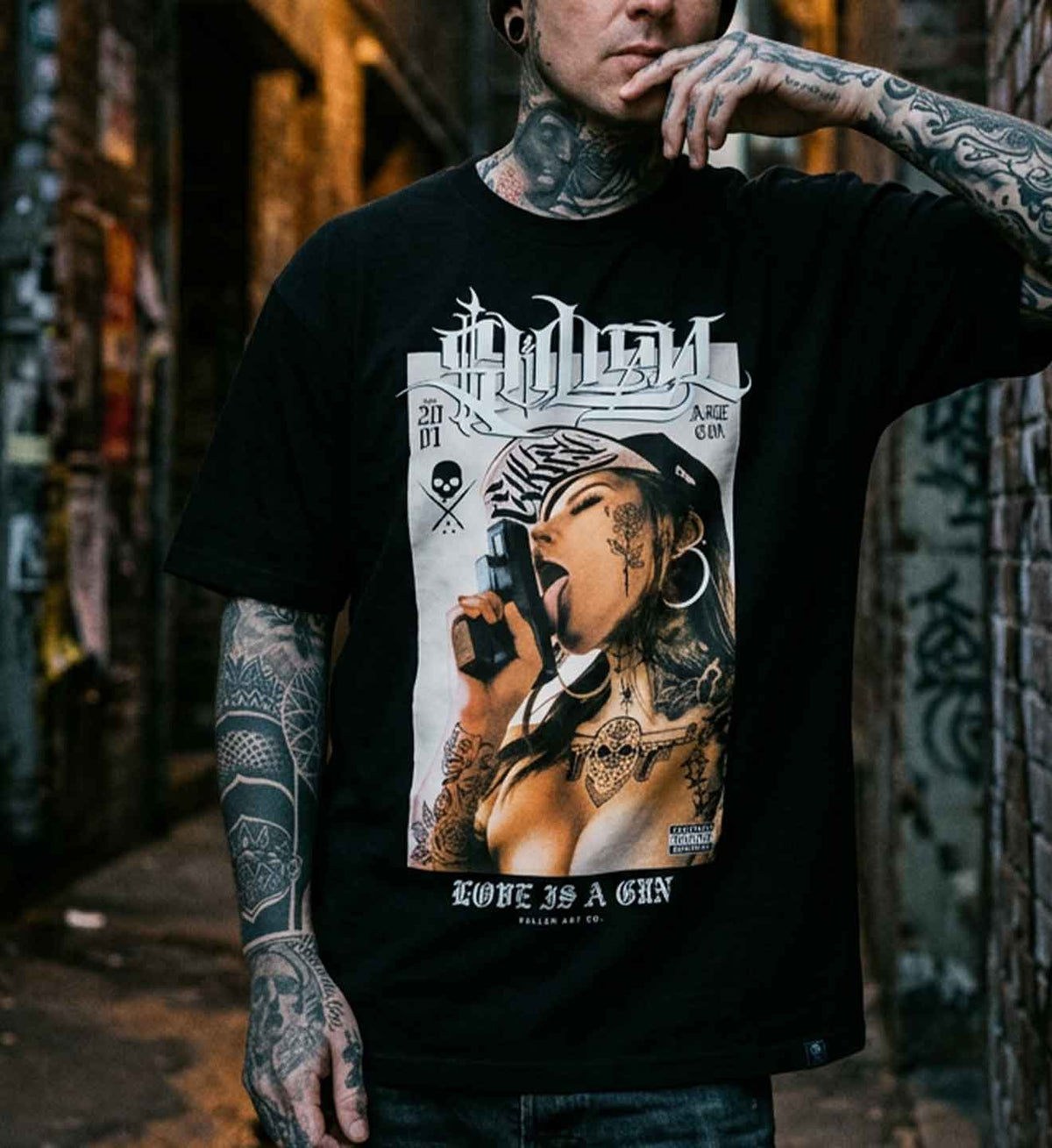 A tattooed man wearing a black graphic t-shirt with a large front print of a woman holding a gun, standing in a graffiti-lined alleyway.