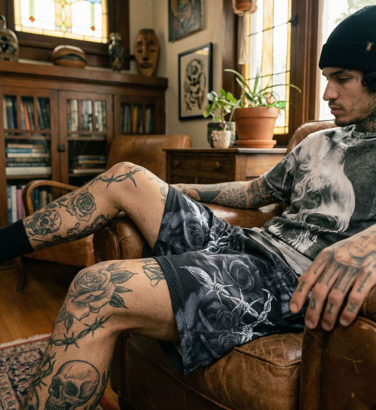 Man with tattoos sitting on a leather couch in a room with bookshelves and plants.
