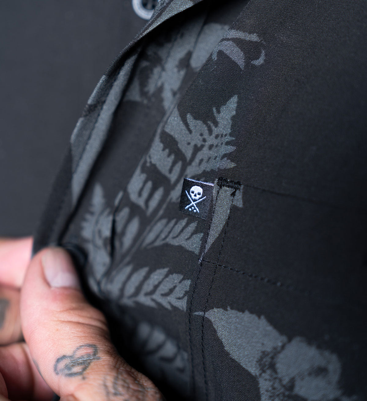 Black shirt with floral pattern and skull emblem held by a person with tattoos.