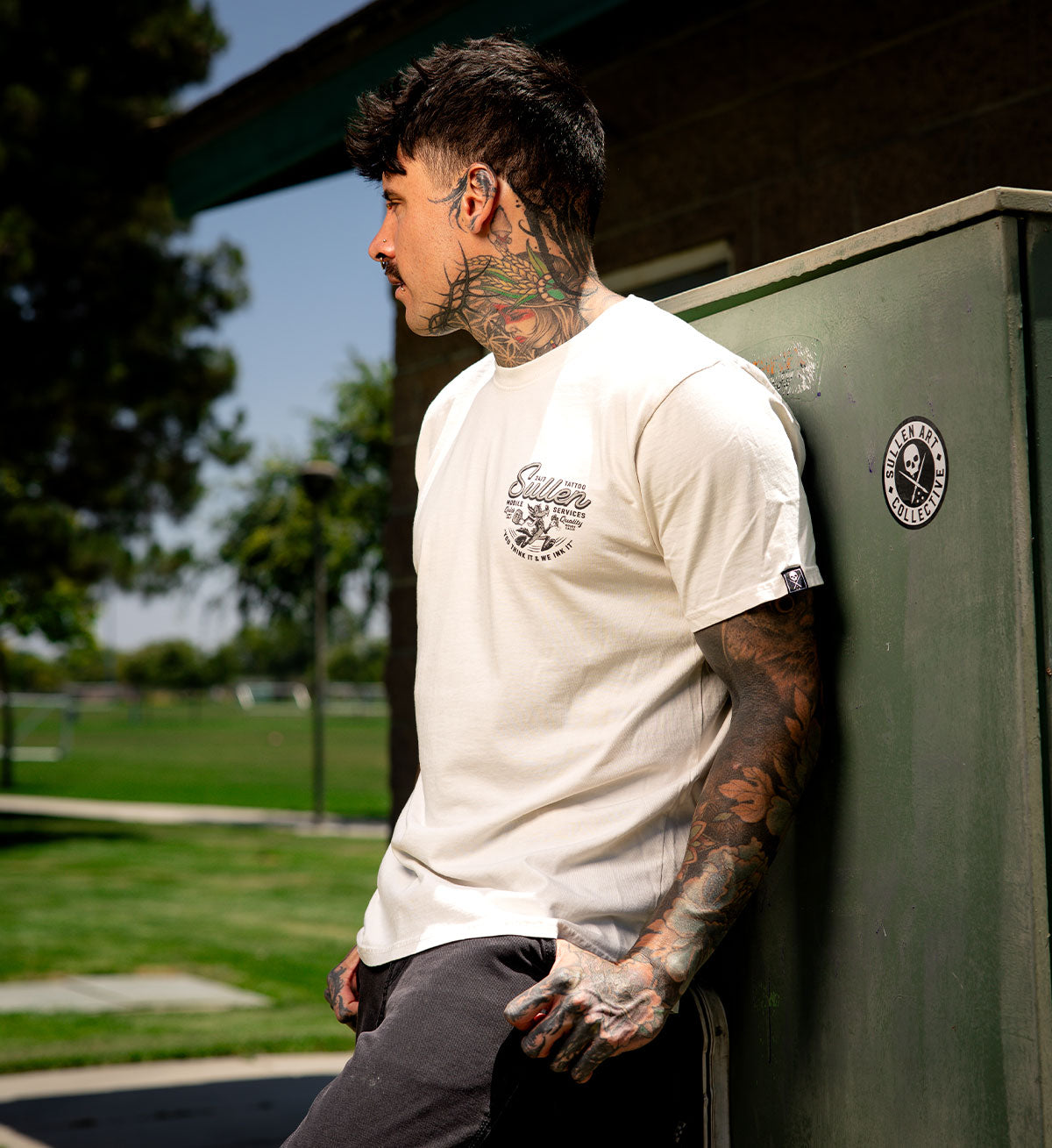 Man with tattoos wearing a white t-shirt leaning against a green wall outdoors.