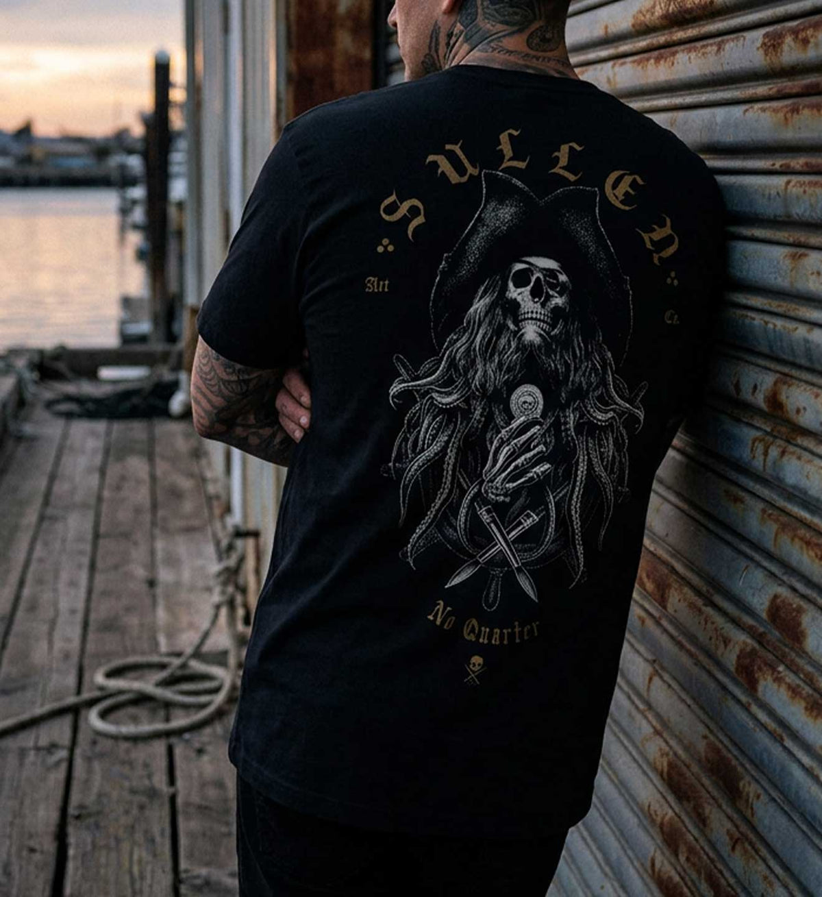 A man stands with his back to the camera wearing a black graphic t-shirt featuring a large gothic skeleton holding a microphone, with the text 'Sullen' and 'A Quarter' on the back, set against a rustic dock and corrugated metal wall background. 2026 Sullen Pirate Day capsule short sleeve tee