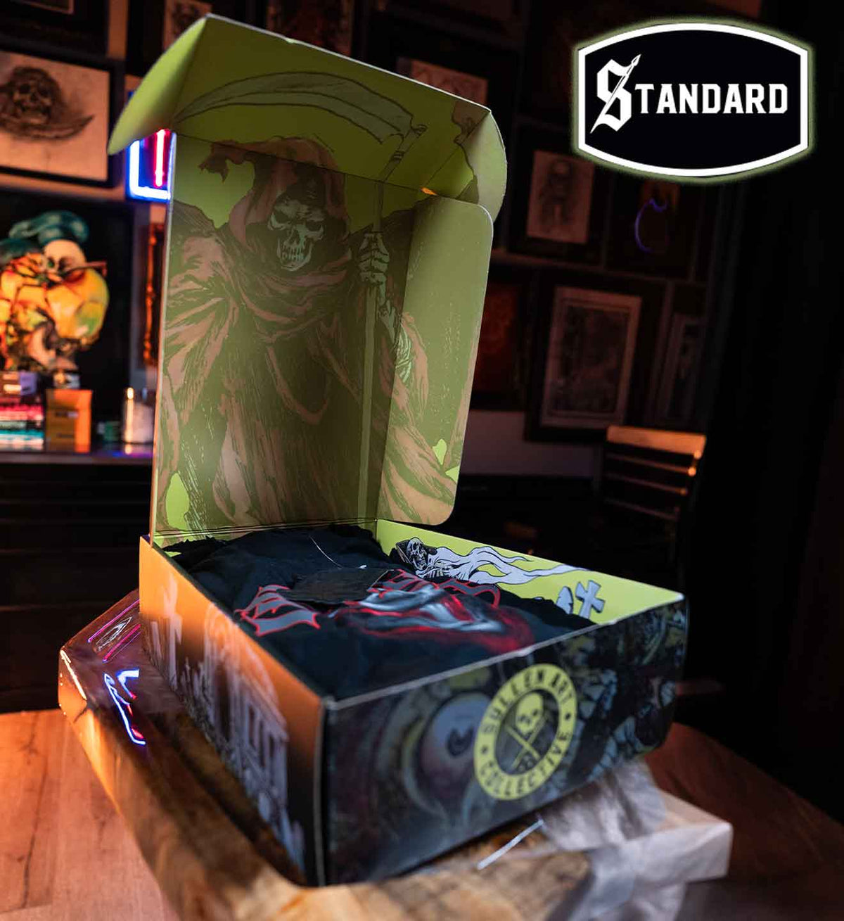 A branded Standard brand box set displayed on a wooden table in a dimly lit room. The open box features a grim reaper design on the inside lid and a dark, illustrated exterior with a circular 'Standard' logo on the front.