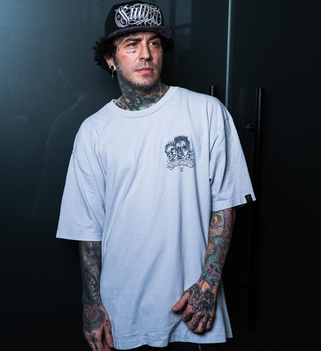 Person wearing a light gray t-shirt with a small logo and a black cap with white text against a dark background.