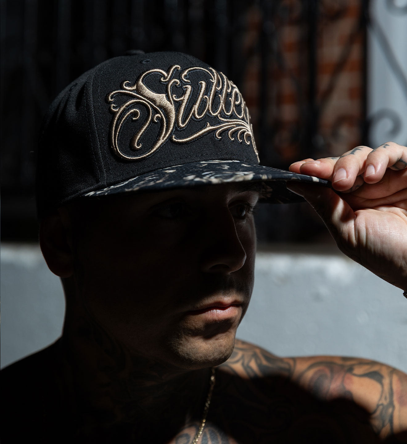 Person wearing a black cap with gold embroidery, holding it with one hand.