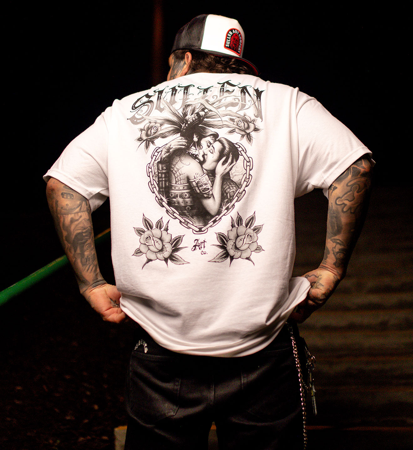 Person wearing a white t-shirt with a graphic design on the back, standing against a dark background.