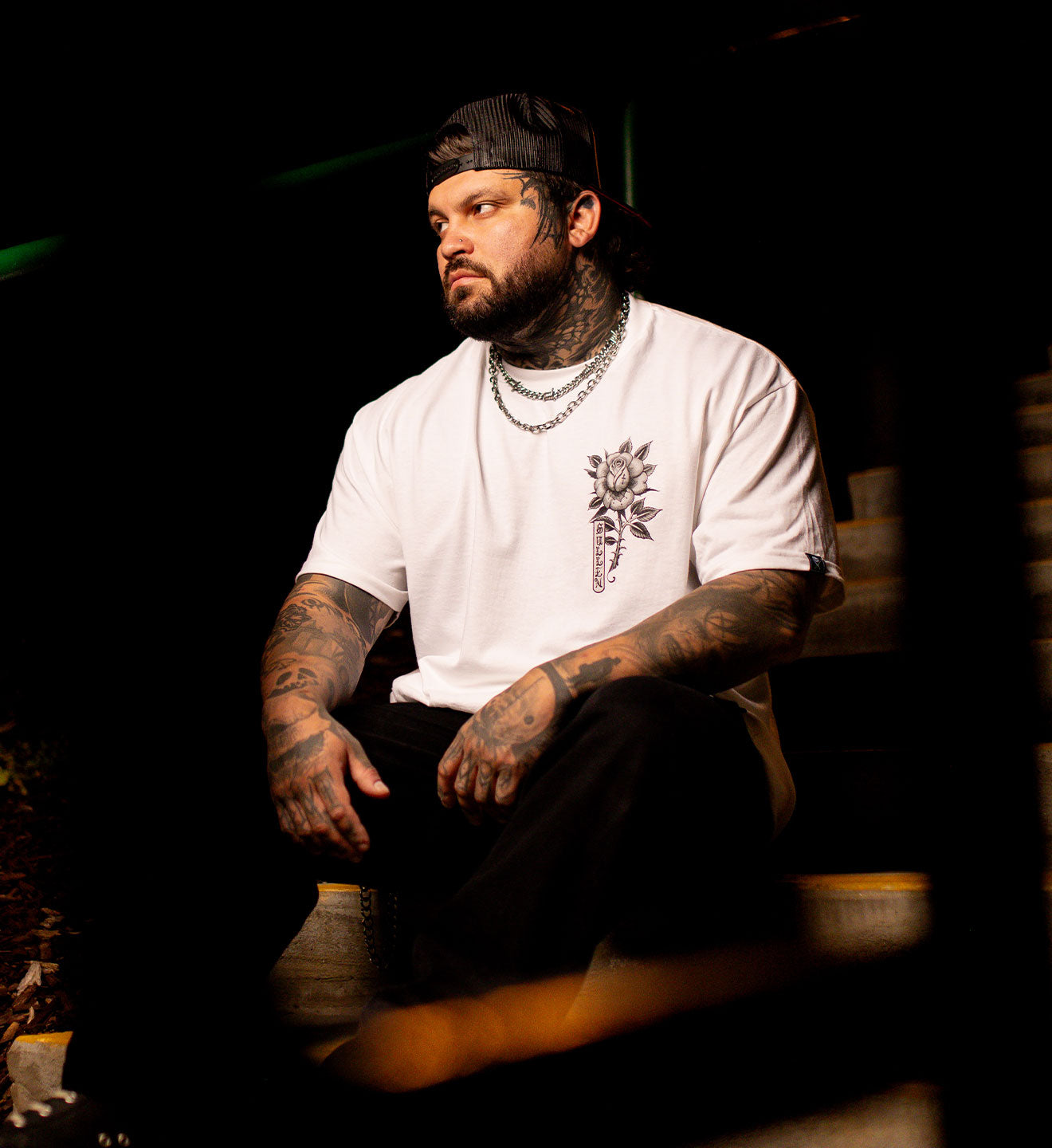 Man with tattoos wearing a white t-shirt and black cap sitting on a dark background