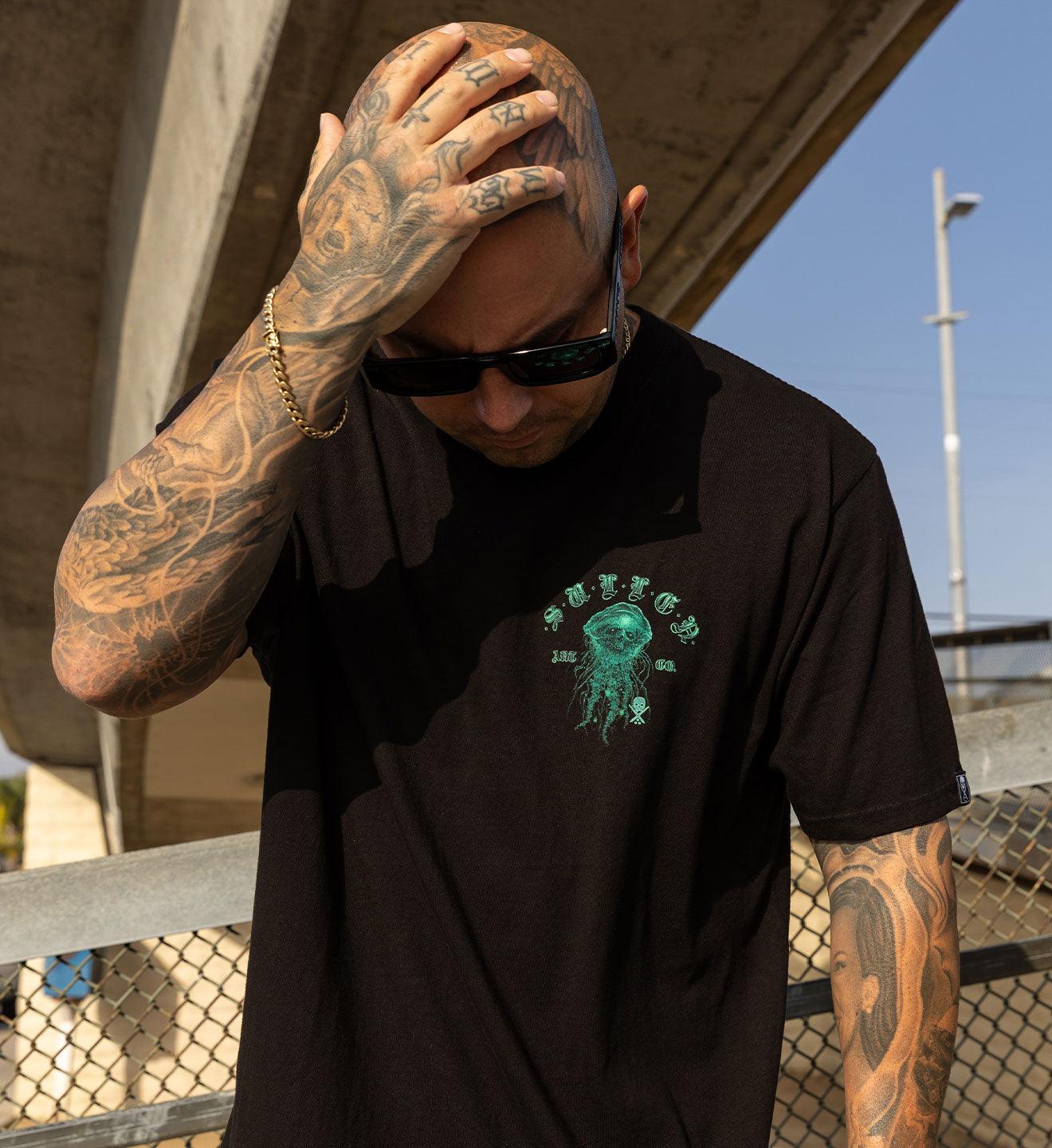 Man with tattoos wearing a black t-shirt with a jellyfish graphic, standing outdoors.