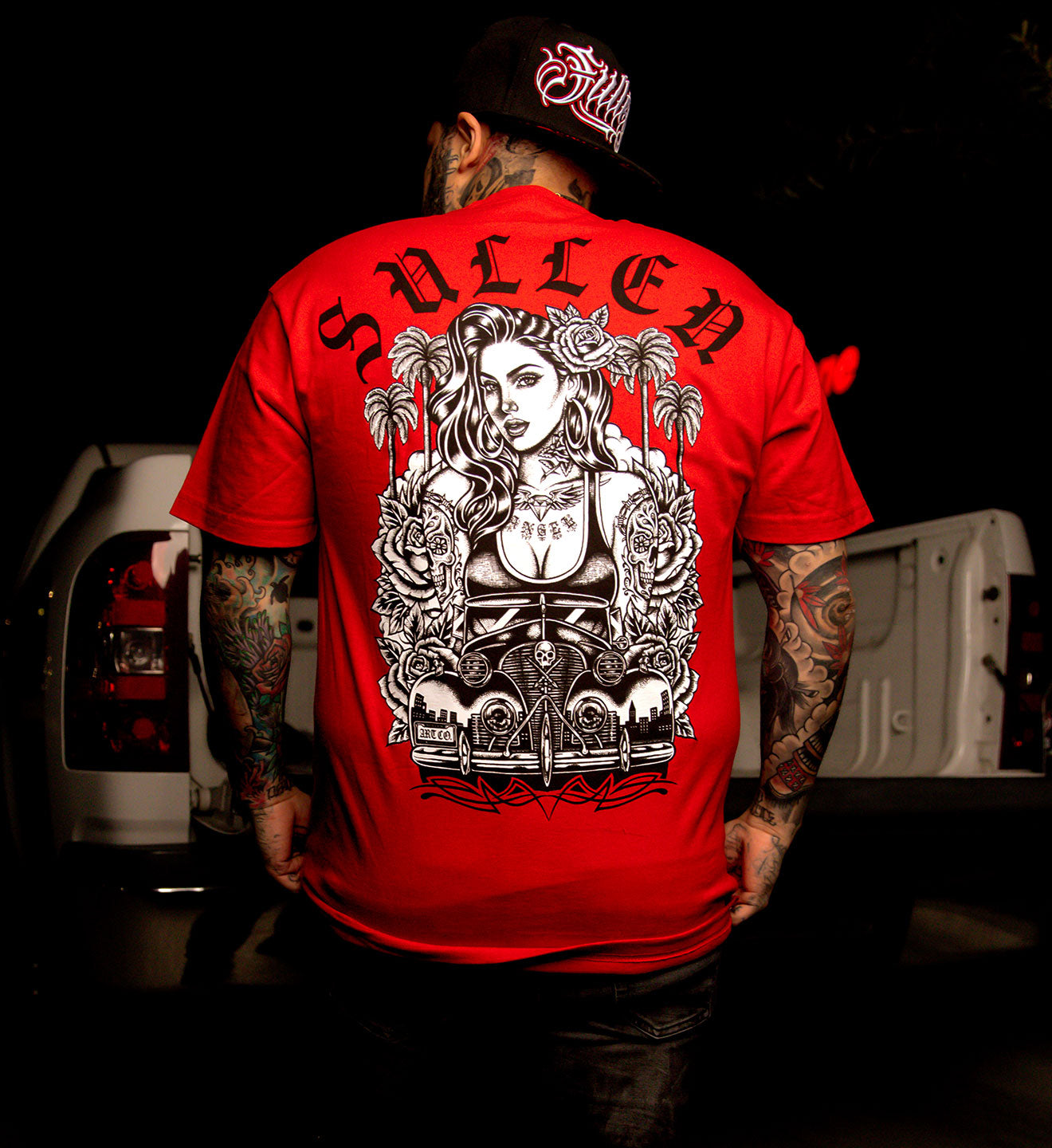 Person wearing a red 'Sullen' t-shirt with a graphic design on the back, standing in front of a vehicle.