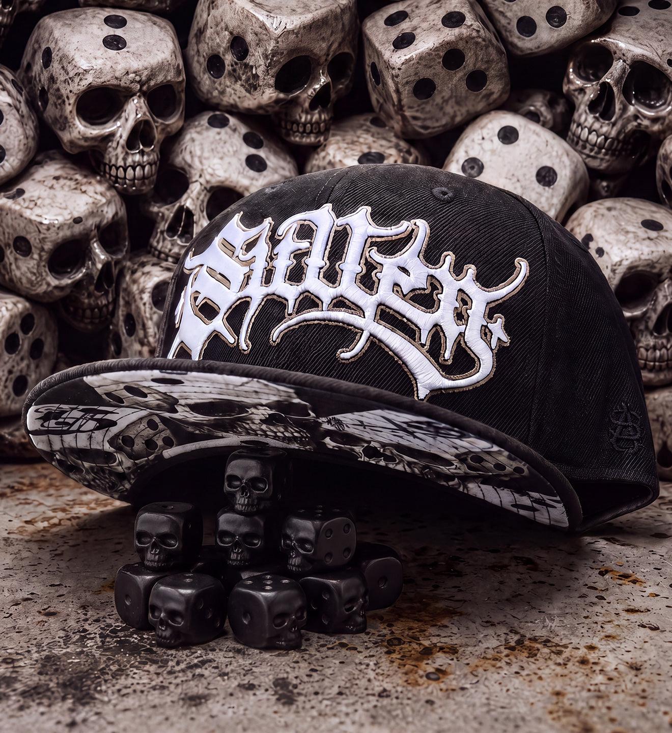 A black snapback cap with a large white gothic-style logo on the front and a skull-and-crossbones pattern on the brim, displayed in front of a pile of skull-shaped dice.
