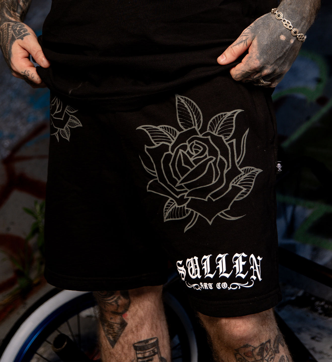 Black shorts with white rose design and 'Sullen' branding worn by a person with tattoos.