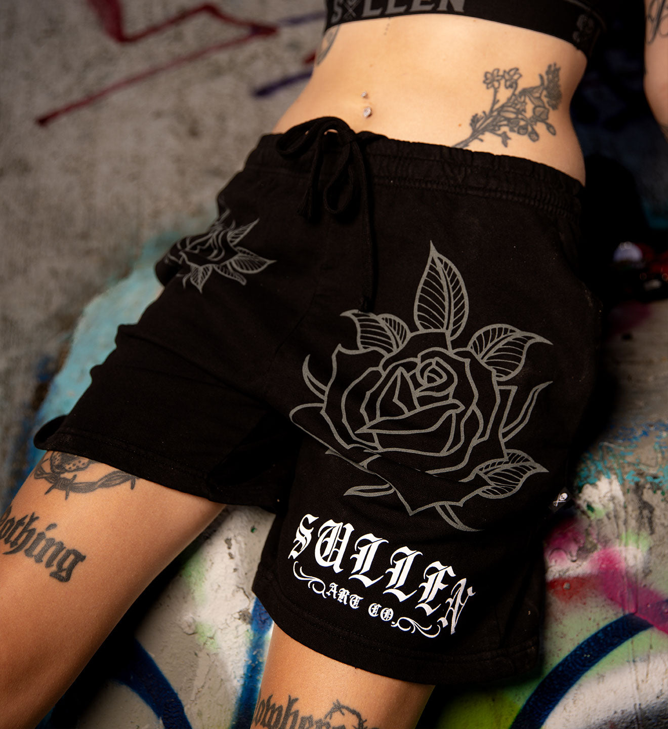 Black shorts with rose design and 'Sullen Art Co' branding worn by a person with tattoos.