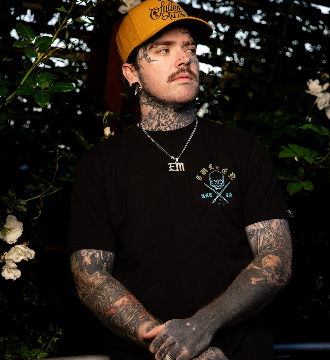 Man with tattoos wearing a black t-shirt with a logo and a yellow cap, sitting outdoors.
