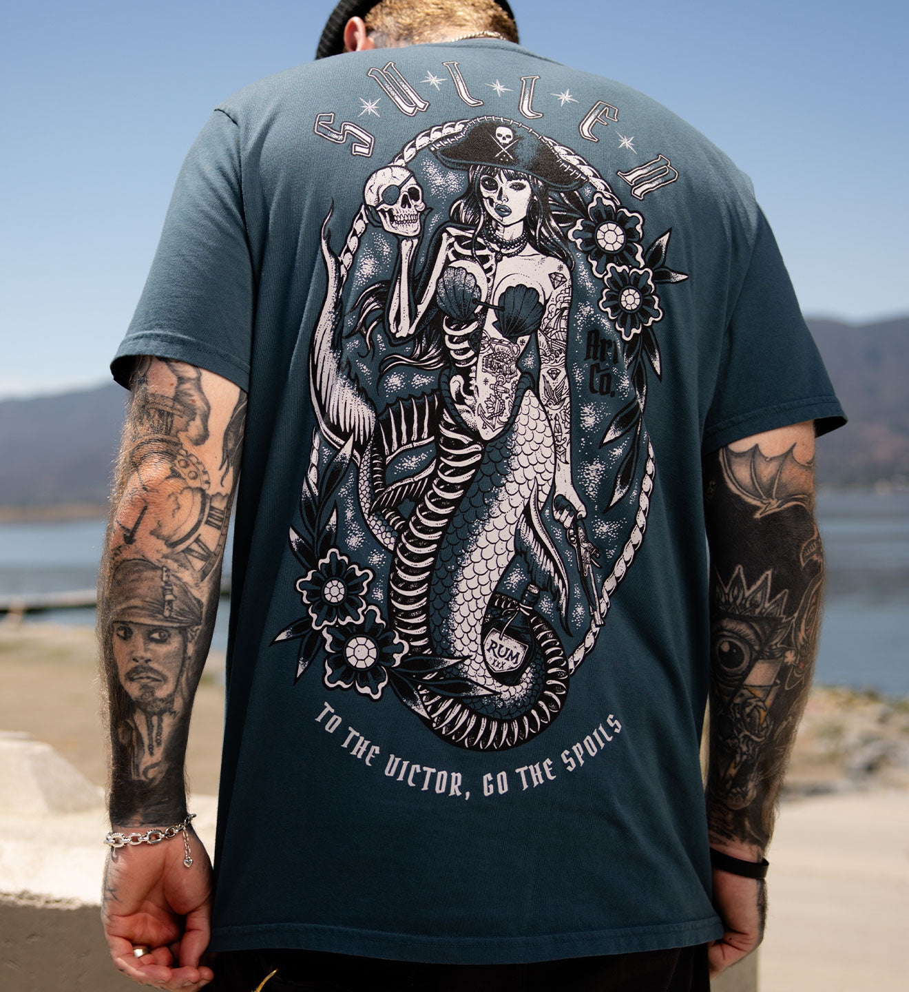 Person wearing a t-shirt with a mermaid and skeleton design on the back, standing outdoors.