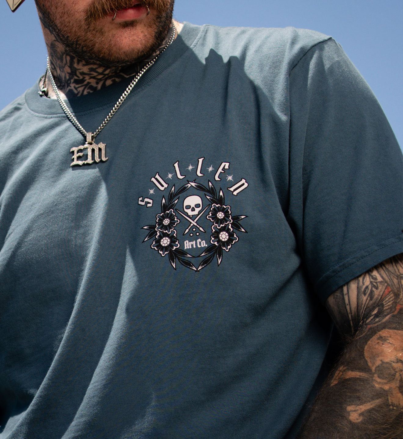 Man wearing a blue t-shirt with a skull and floral design, standing against a clear blue sky.