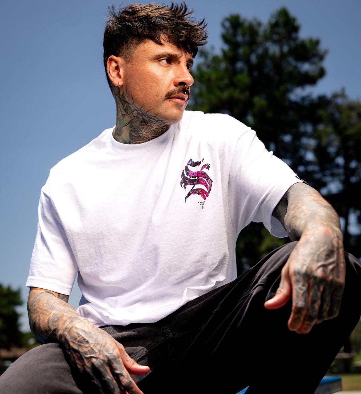 Man wearing a white t-shirt with a colorful logo, sitting outdoors with trees in the background.