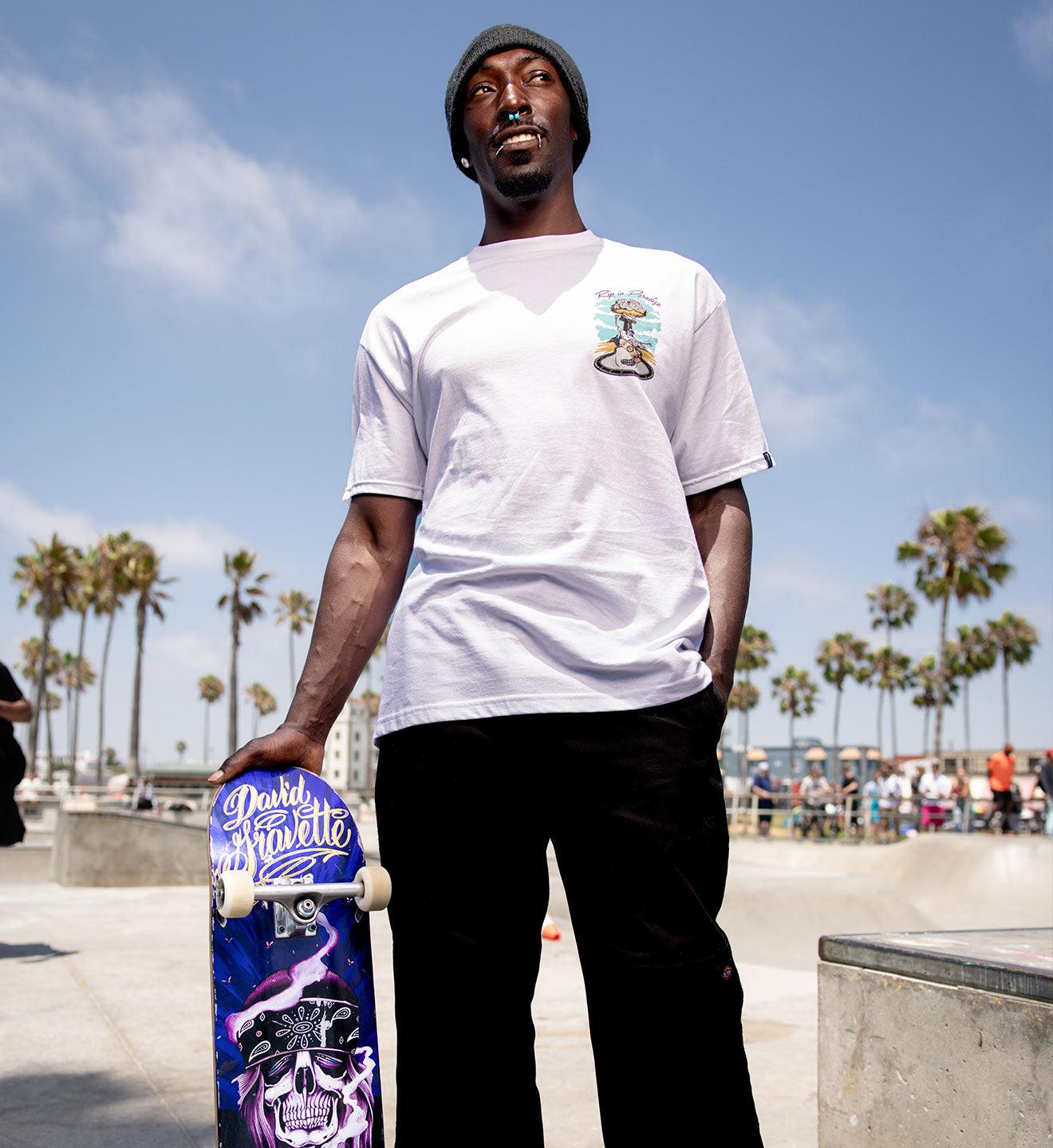 Man holding a skateboard at a skate park with palm trees in the background