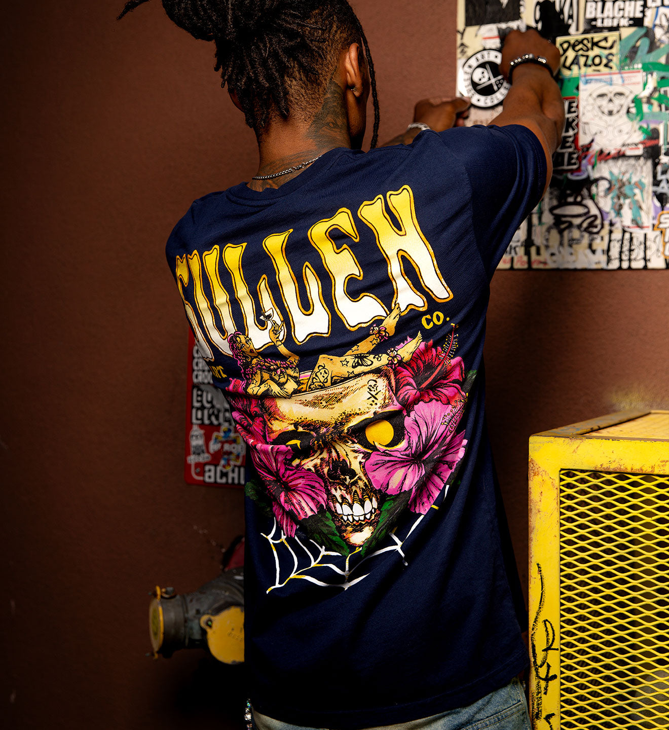 Person wearing a dark blue t-shirt with a colorful graphic design on the back, standing against a brown wall.